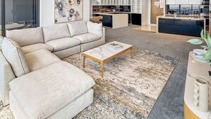 A modern living room with a sofa, coffee table, and a rug. There is a kitchen in the background with cabinets and appliances.