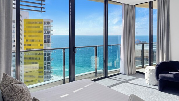 A modern hotel room with a large balcony, a bed, a chair, and a view of the ocean and buildings.