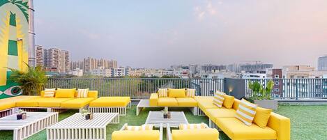 A rooftop terrace with yellow and white furniture, a view of the cityscape, and a mural on the left.