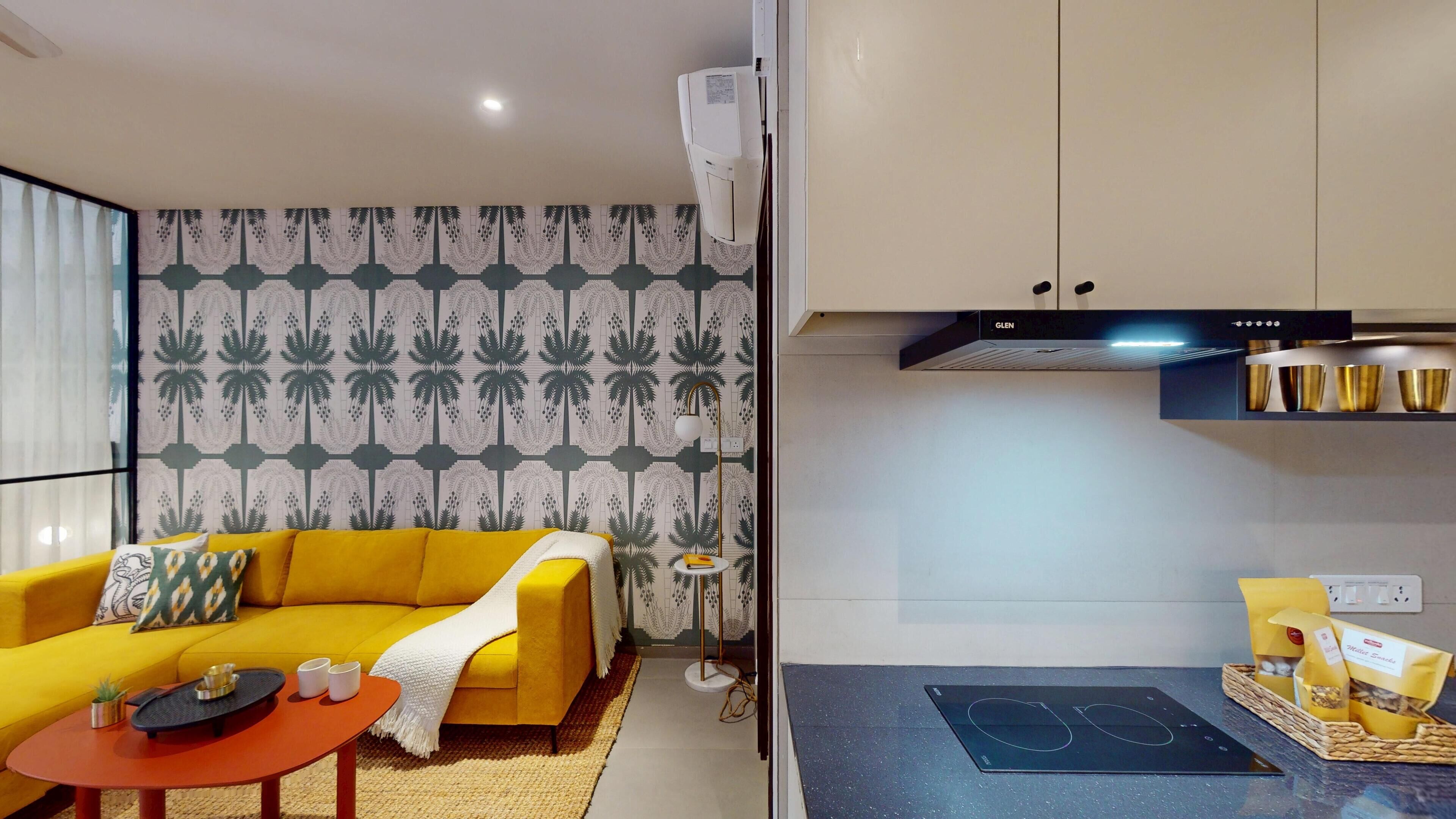A compact living space with a yellow sofa, a small coffee table, a modern kitchen with a built-in oven, and a patterned wallpaper.