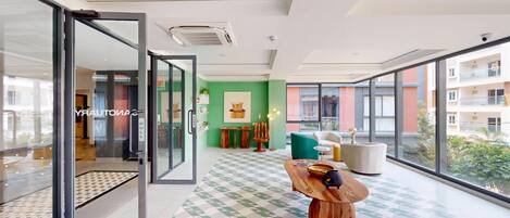 Un moderno vestíbulo de hotel con entrada por puerta de vidrio, una pared verde con obras de arte, una mesa de centro de madera con objetos decorativos y vistas a la ciudad a través de amplios ventanales.