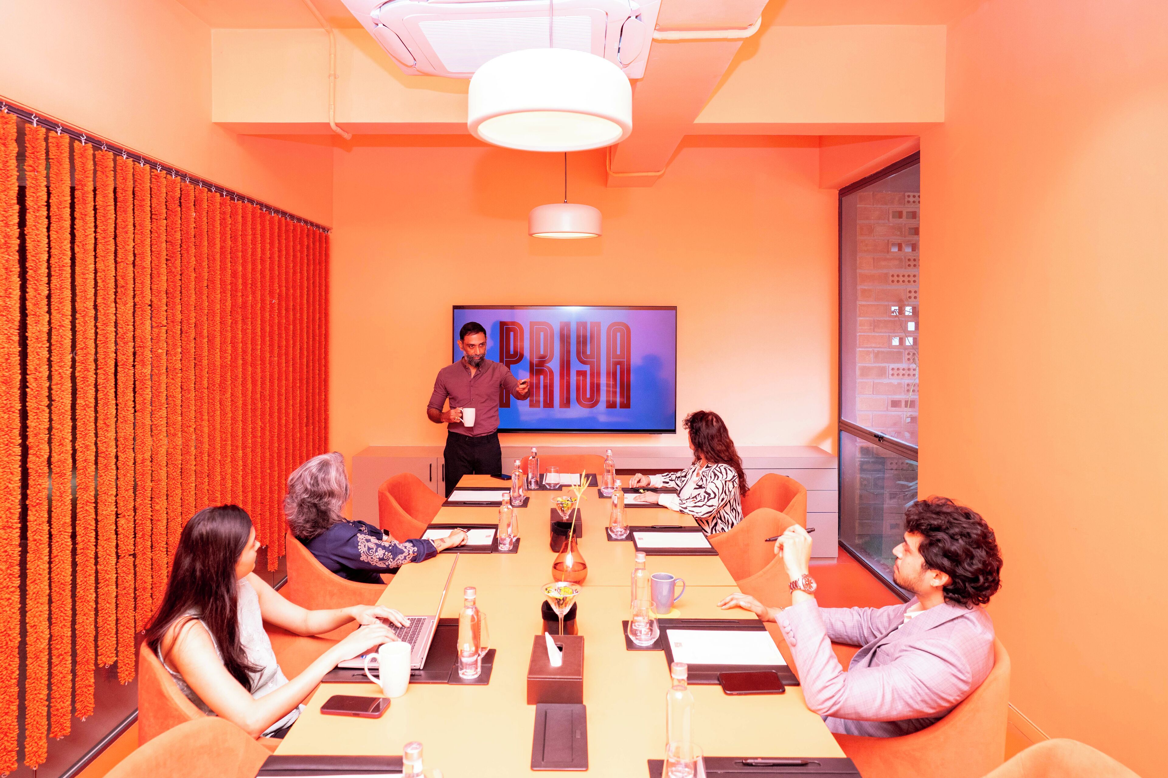 A conference room with a large screen displaying the word "PRYA" and a man presenting.