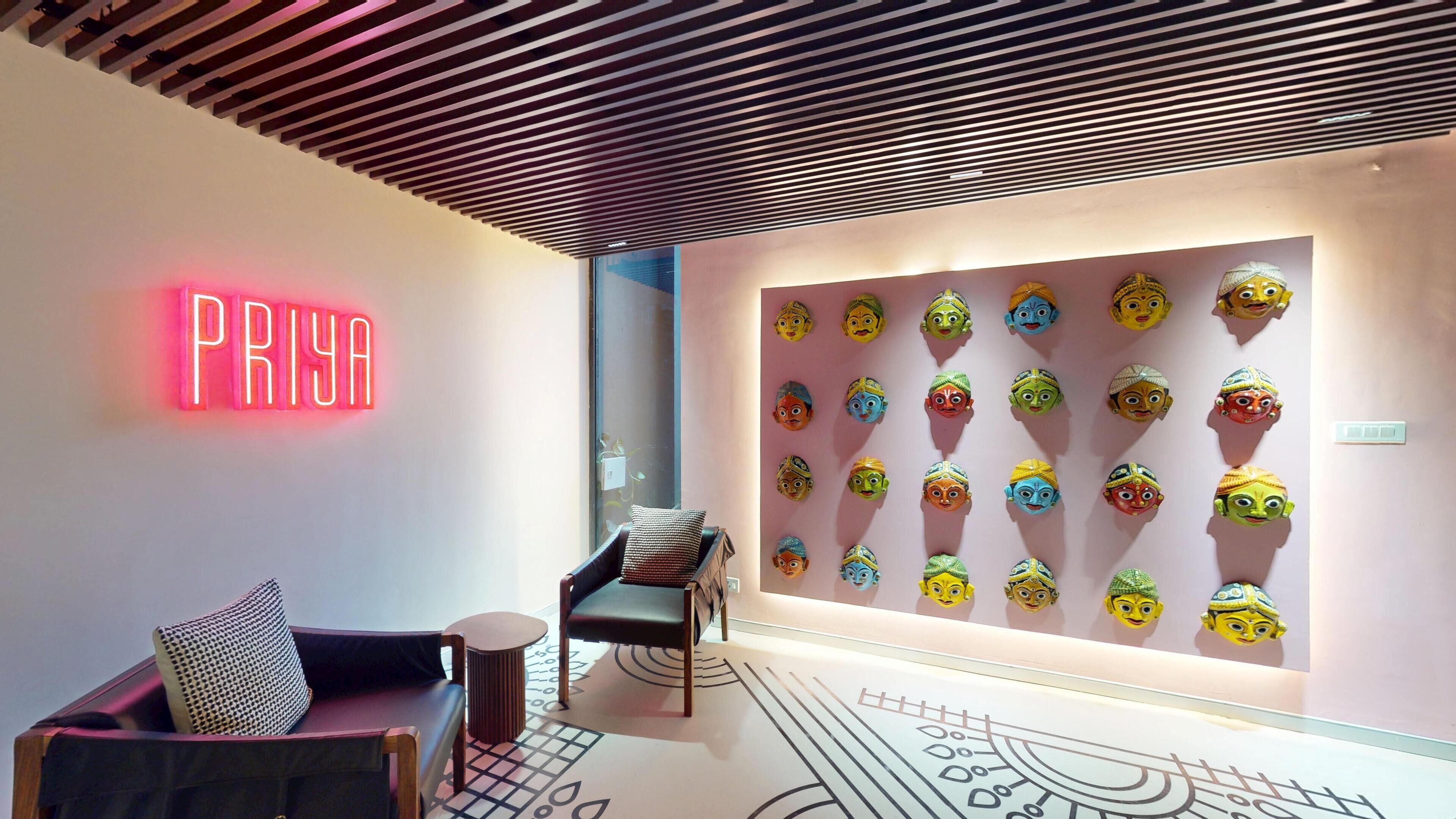 A modern interior with a neon sign reading "PRIYA," a wall of colorful masks, a patterned floor, and a wooden ceiling.