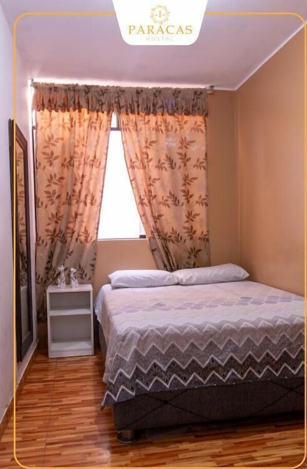 A bedroom with a bed, a nightstand, and a window with curtains. - Hostal Paracas I (Ica)