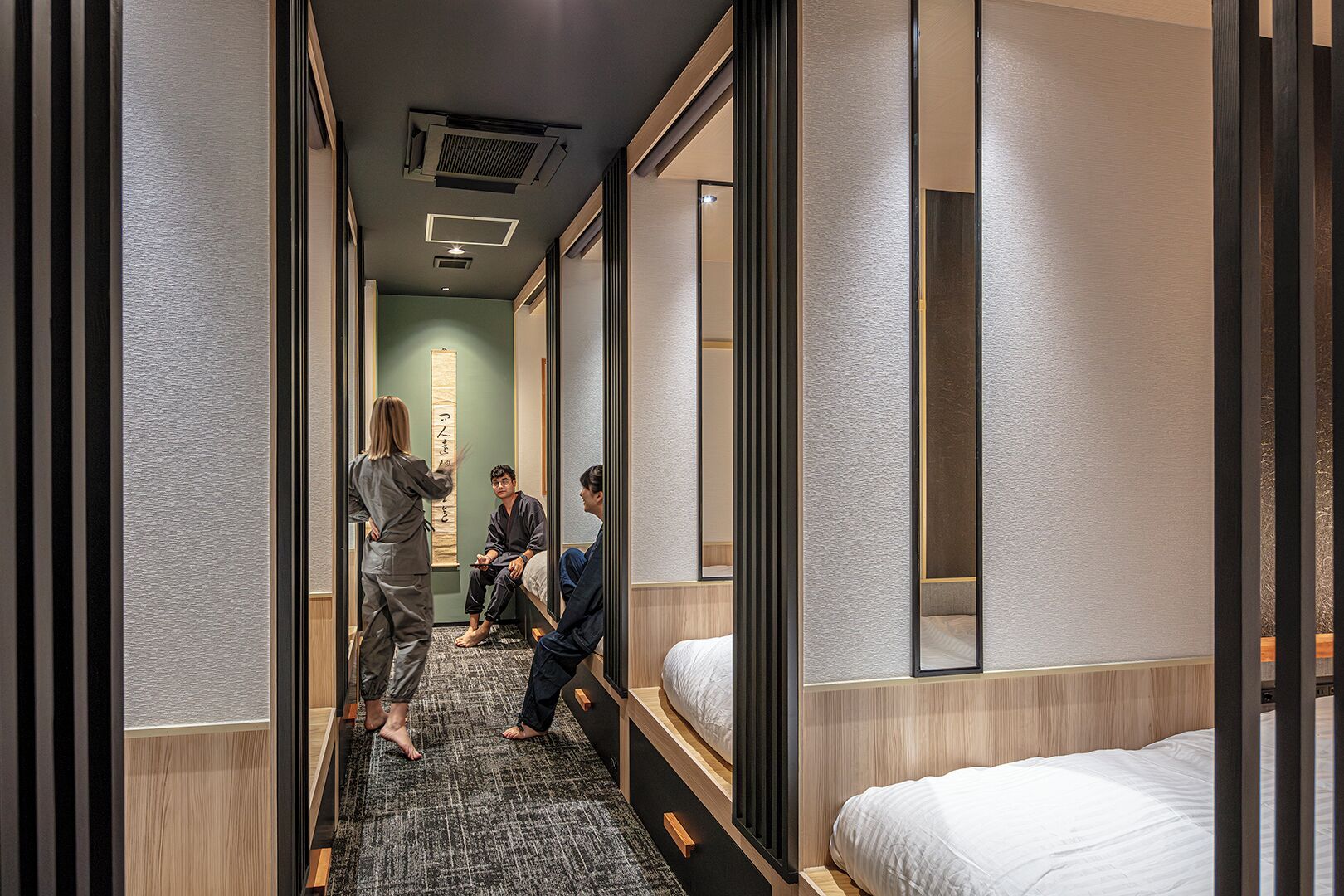 A narrow hotel corridor with a wooden floor, a bed on the right, and a person standing near the bed.