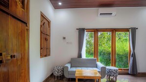 Un salon avec un canapé gris, une table basse en bois et des poufs à motifs. Une armoire en bois et un tableau ornent le mur de gauche. De grandes fenêtres, dotées de rideaux, offrent une vue sur la verdure à l’extérieur.