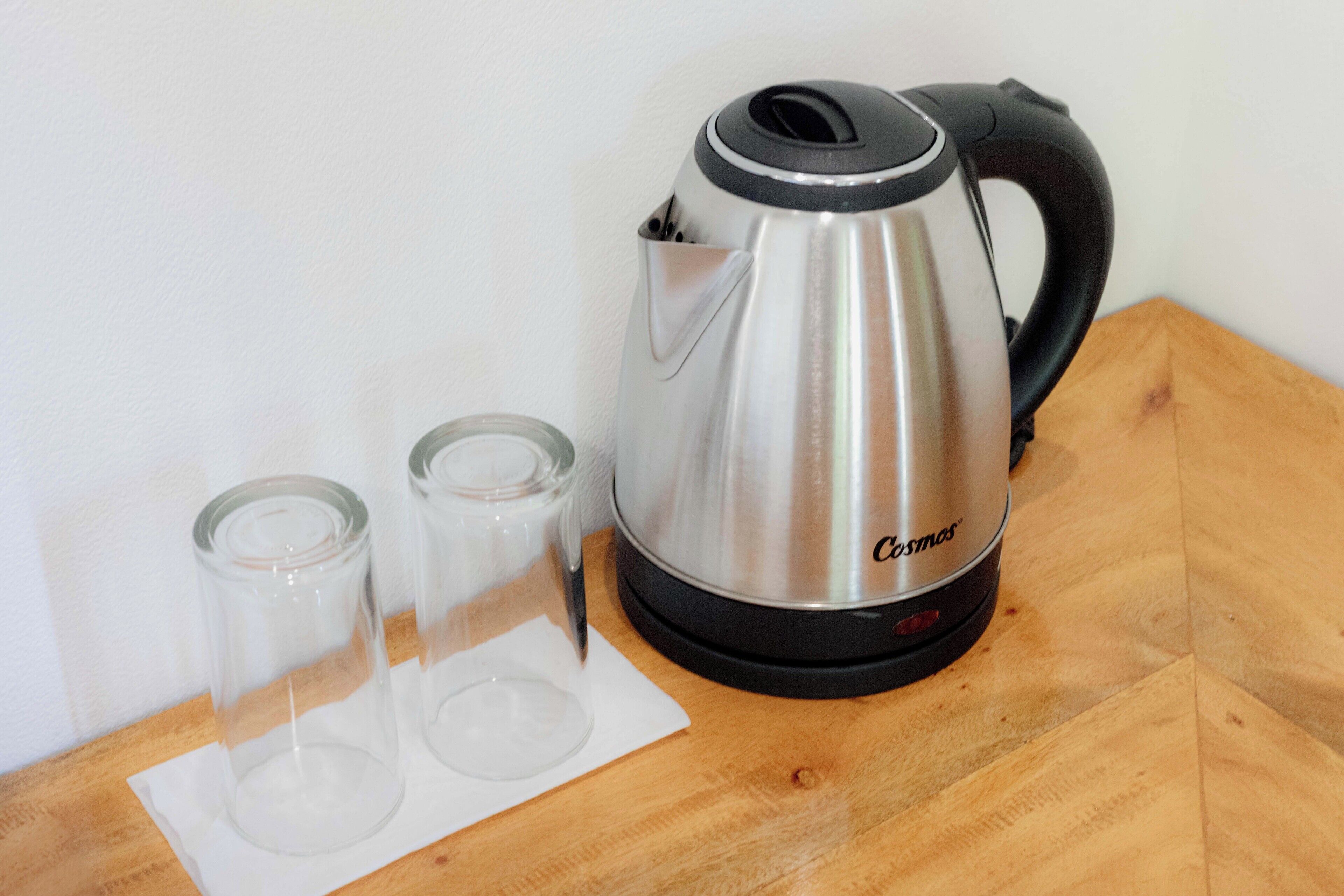 Uma chaleira elétrica Cosmos de aço inoxidável sobre uma mesa de madeira, com duas chávenas de vidro vazias e um guardanapo branco.