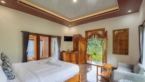 Une chambre dotée d’un grand lit, d’une armoire en bois, d’une télévision et d’un balcon offrant une vue sur la verdure.