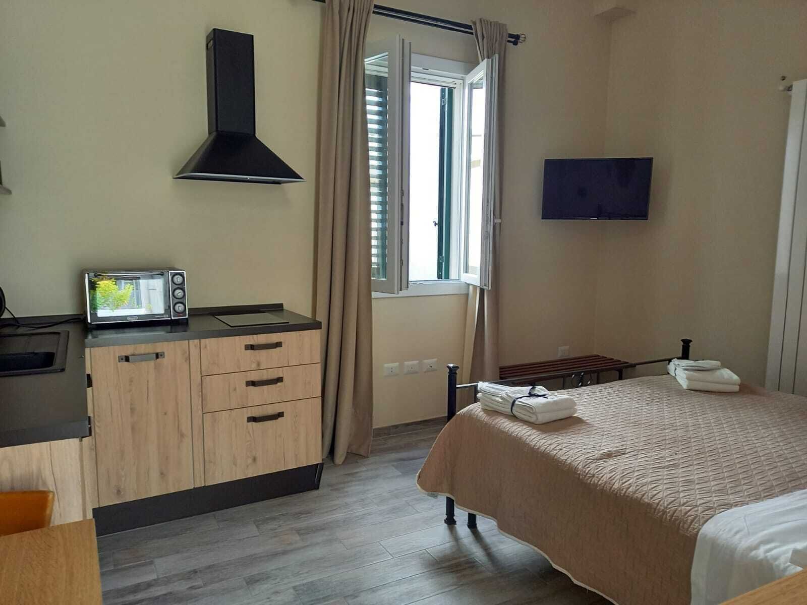 Een hotelkamer met kitchenette, een bed met een beige dekbedovertrek, een aan de muur bevestigde televisie en een raam met gordijnen.