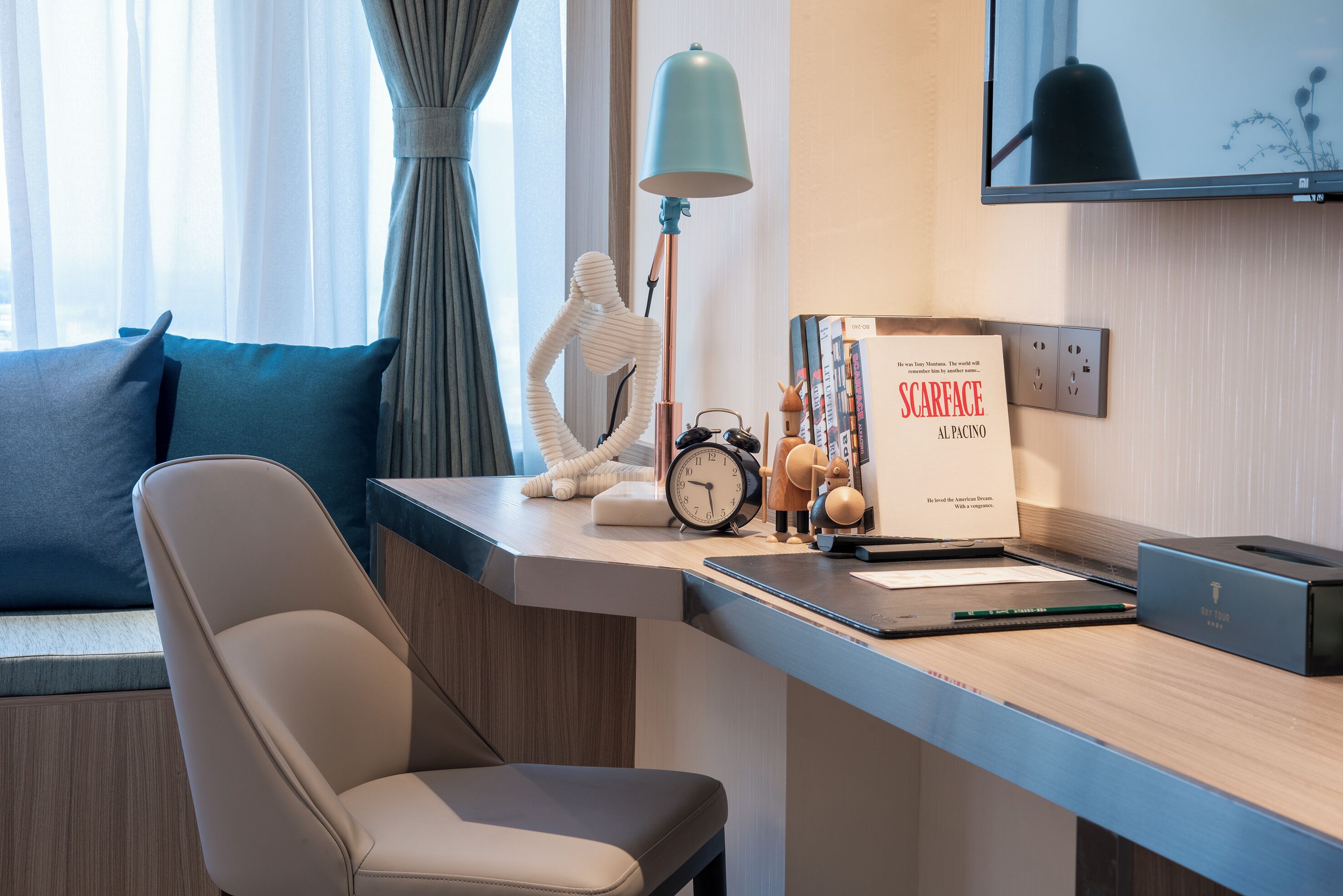 A modern hotel room with a desk, a chair, a lamp, a clock, a decorative figure, and a book titled "SCARFACE AL PACINO".