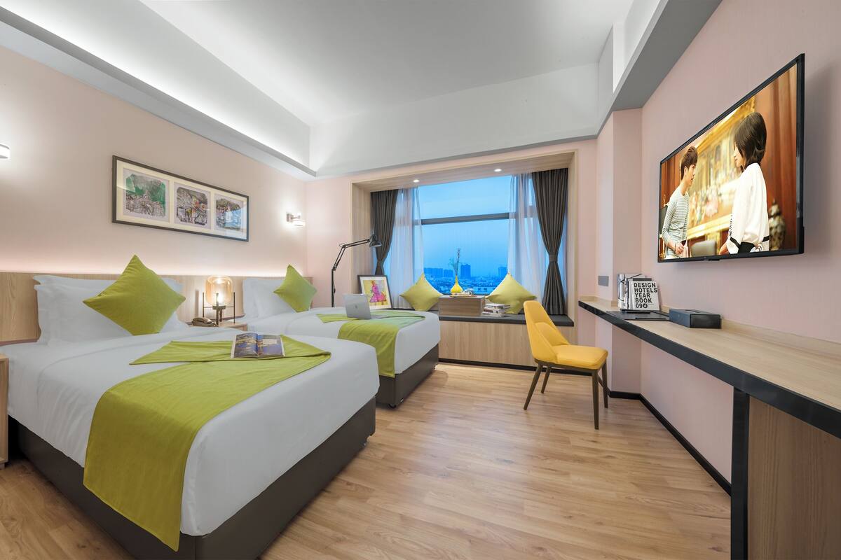 A hotel room with two beds, a desk, a chair, a TV, and a window with a city view.