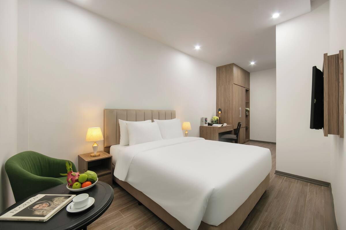 Une chambre d’hôtel avec un grand lit, un fauteuil vert, une table de chevet avec une lampe, un panier de fruits et un magazine.