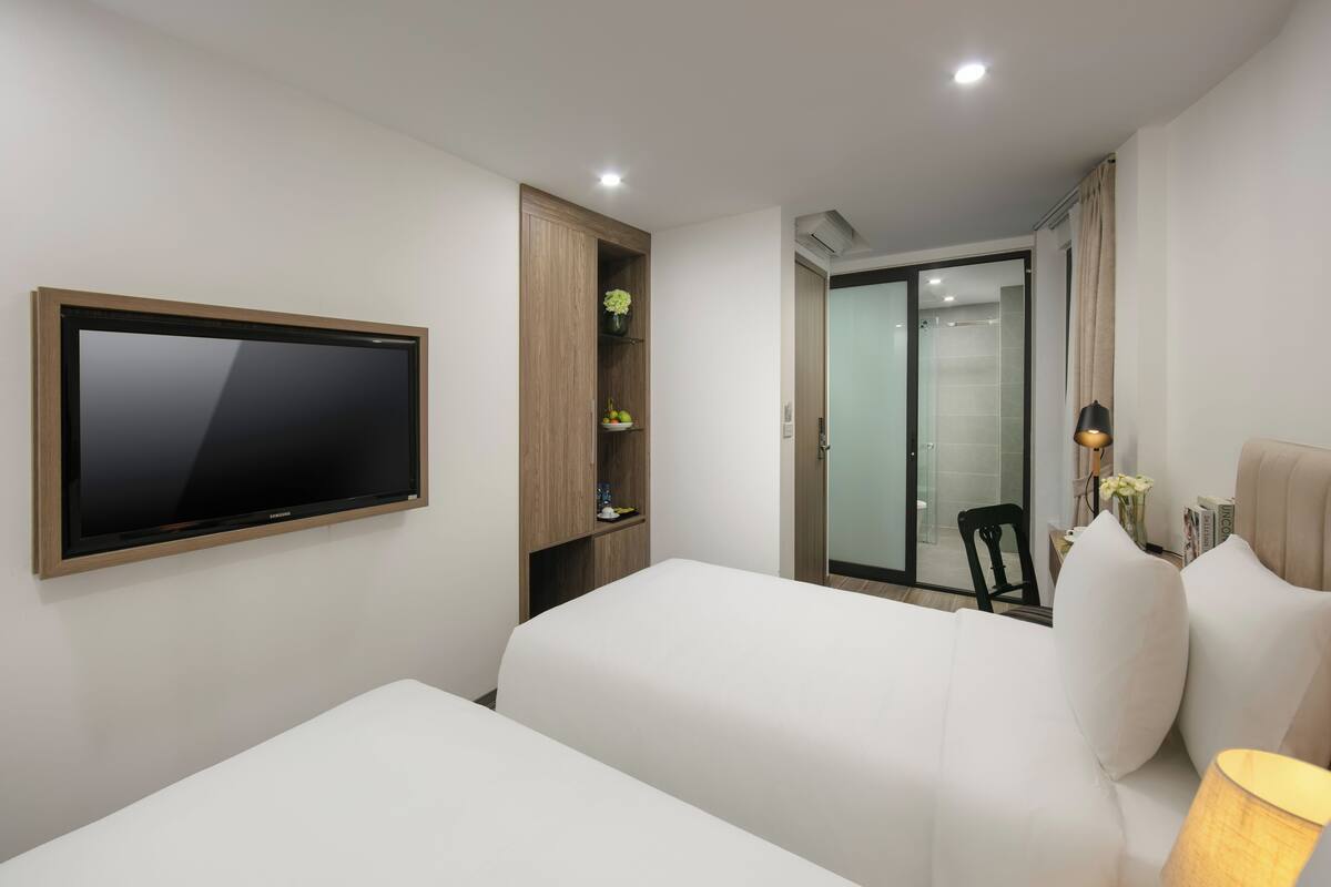 Une chambre d’hôtel dotée d’un téléviseur à écran plat fixé au mur, d’une armoire en bois ornée d’un vase et de fleurs, d’un lit avec une literie blanche, et d’une salle de bain visible à travers une porte en verre.