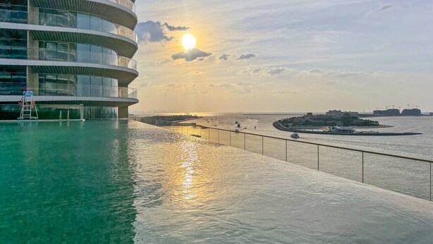 A waterfront hotel with a pool, a sunlit sky, and a distant view of the sea. - Coastal Elegance 1-Bedroom in Grand Bleu (Emaar Beachfront)