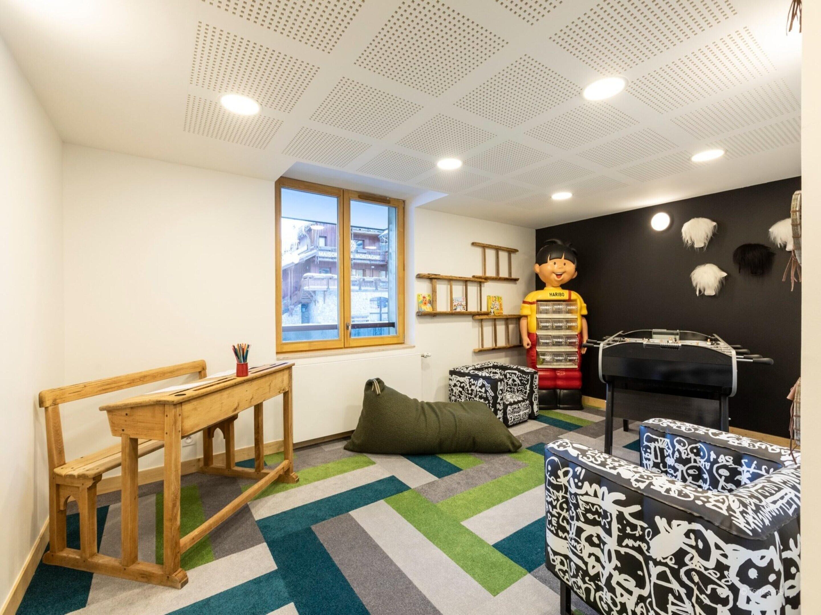 A children's playroom with a large window, a wooden bench, a green bean bag, and a black and white patterned ottoman.