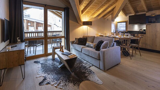 A cozy living room with a sofa, coffee table, and a rug. There is a dining area with a table and chairs. The room has wooden beams on the ceiling and a large window.