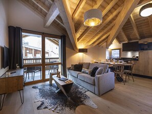 Un accogliente soggiorno con divano, tavolino e tappeto. C’è una zona pranzo con tavolo e sedie. La stanza presenta travi a vista al soffitto e un'ampia finestra.