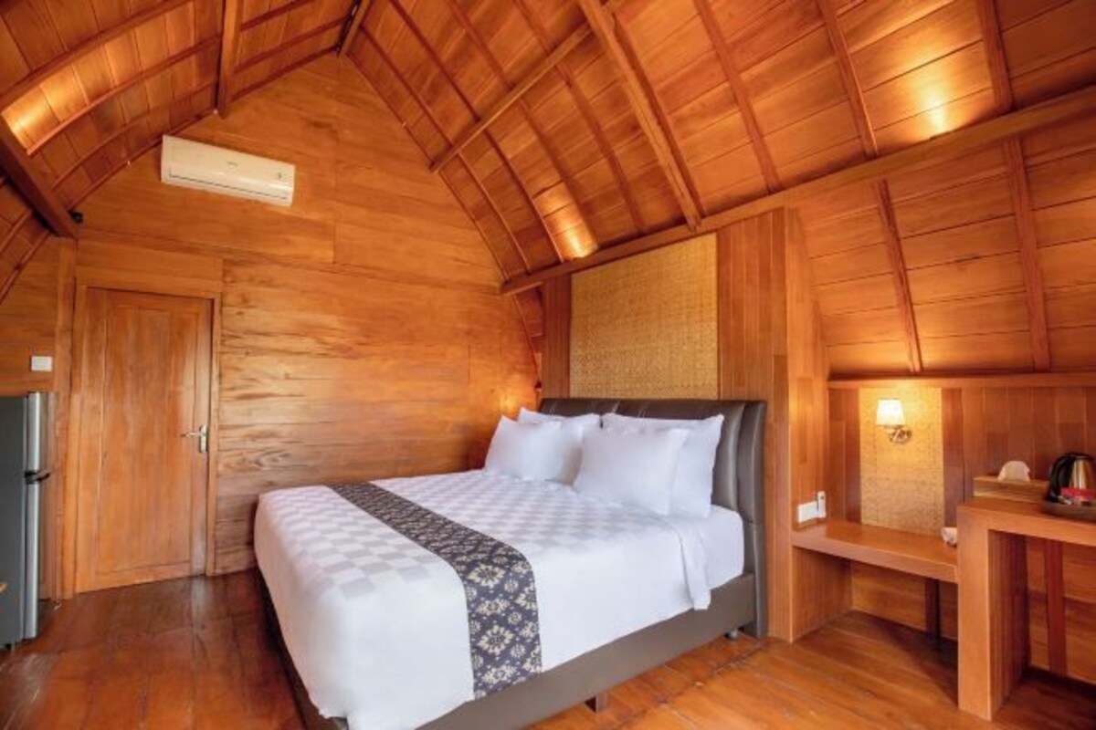 A wooden cabin room with a bed, a small table, and a wall-mounted lamp.