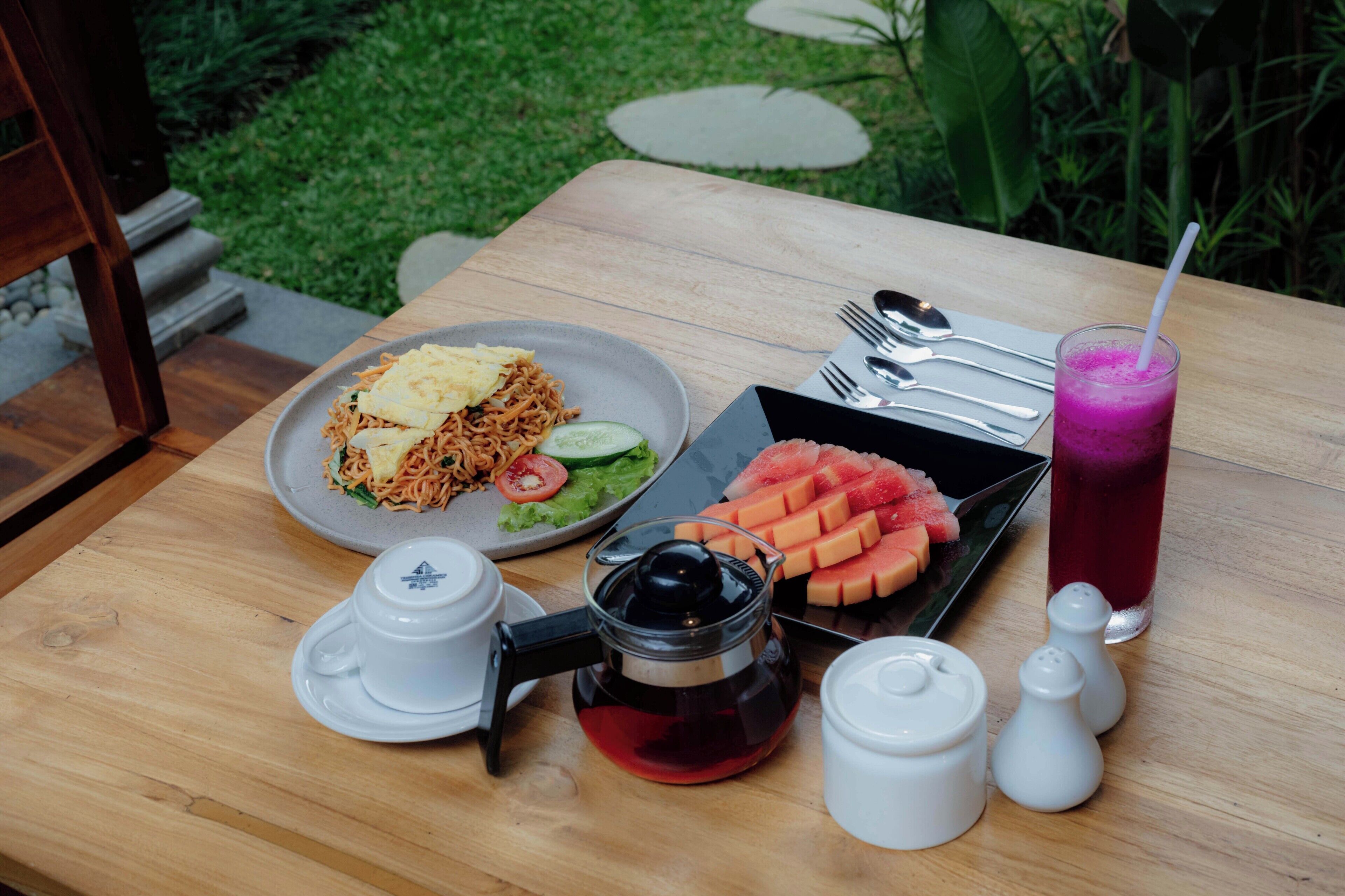 Uma mesa de madeira com uma refeição de macarrão, vegetais e melancia, uma chaleira, saleiro e pimenteiro, e um copo de bebida rosa.