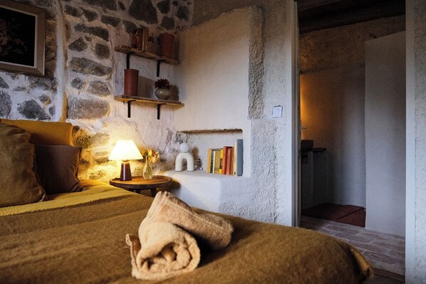 Un dormitori acollidor amb una paret de pedra, una làmpada de nit, una tovallola enrotllada i una estant amb llibres i objectes decoratius.