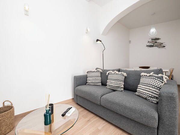 Un salon moderne avec un canapé gris, des coussins à motifs, une table basse en verre, un lampadaire et un luminaire suspendu.