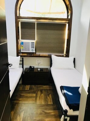 Une chambre de taille modeste, avec deux lits, une petite table et une fenêtre équipée de stores.