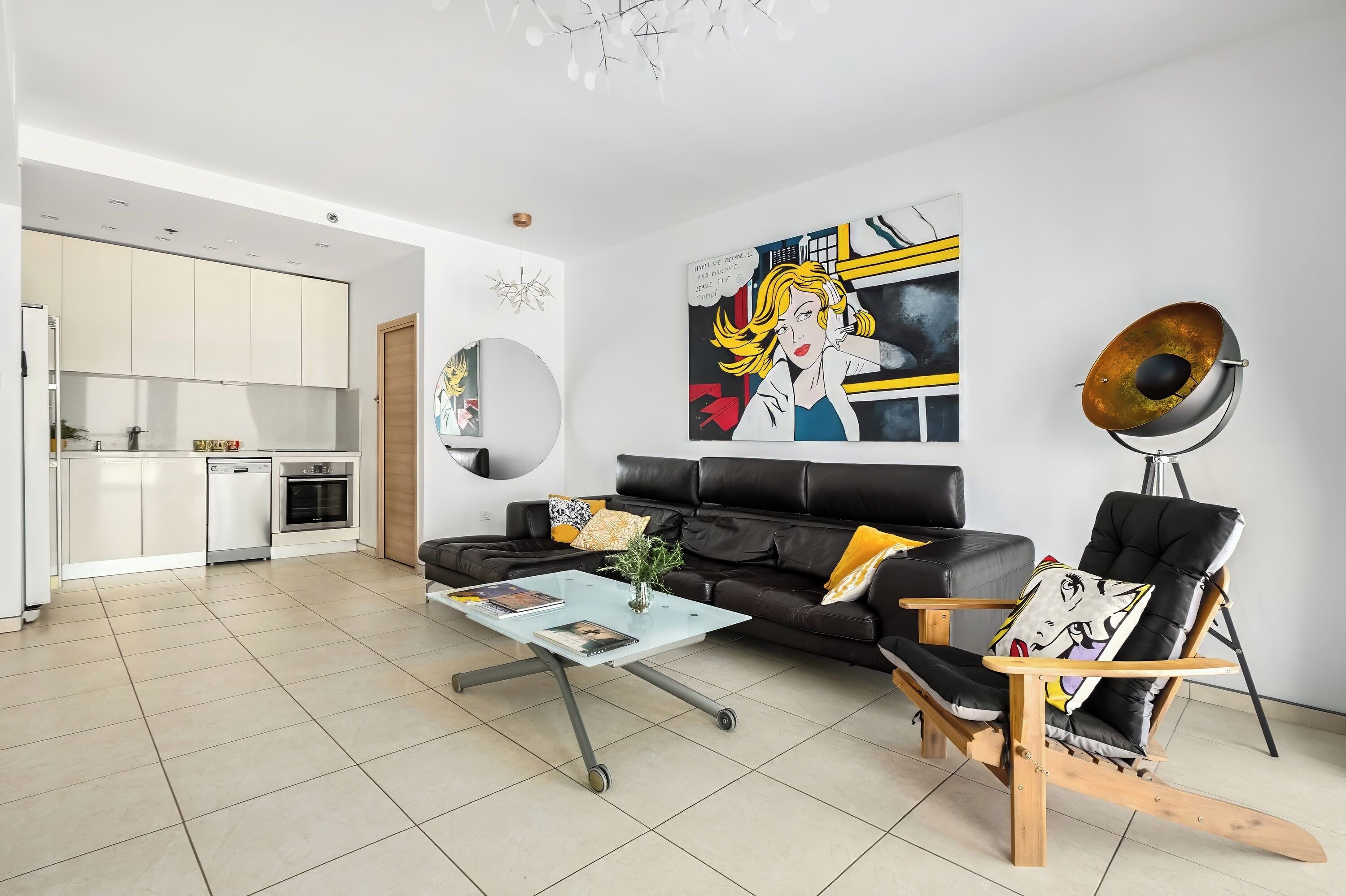 A modern living room with a sofa, a coffee table, and a rocking chair. There is a kitchen in the background with white cabinets and a dishwasher.