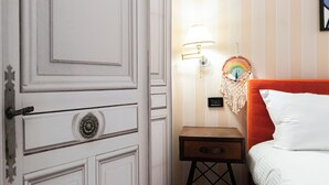 Ett hotellrum med en vit dörr, ett nattduksbord med en lampa, en färgglad väggdekoration och en orange sänggavel.