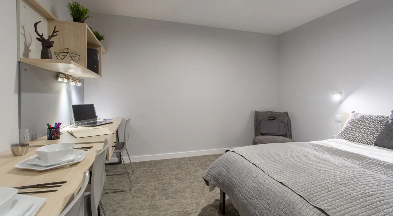 A bedroom with a bed, a desk with a laptop, a chair, and a shelf with decorative items.