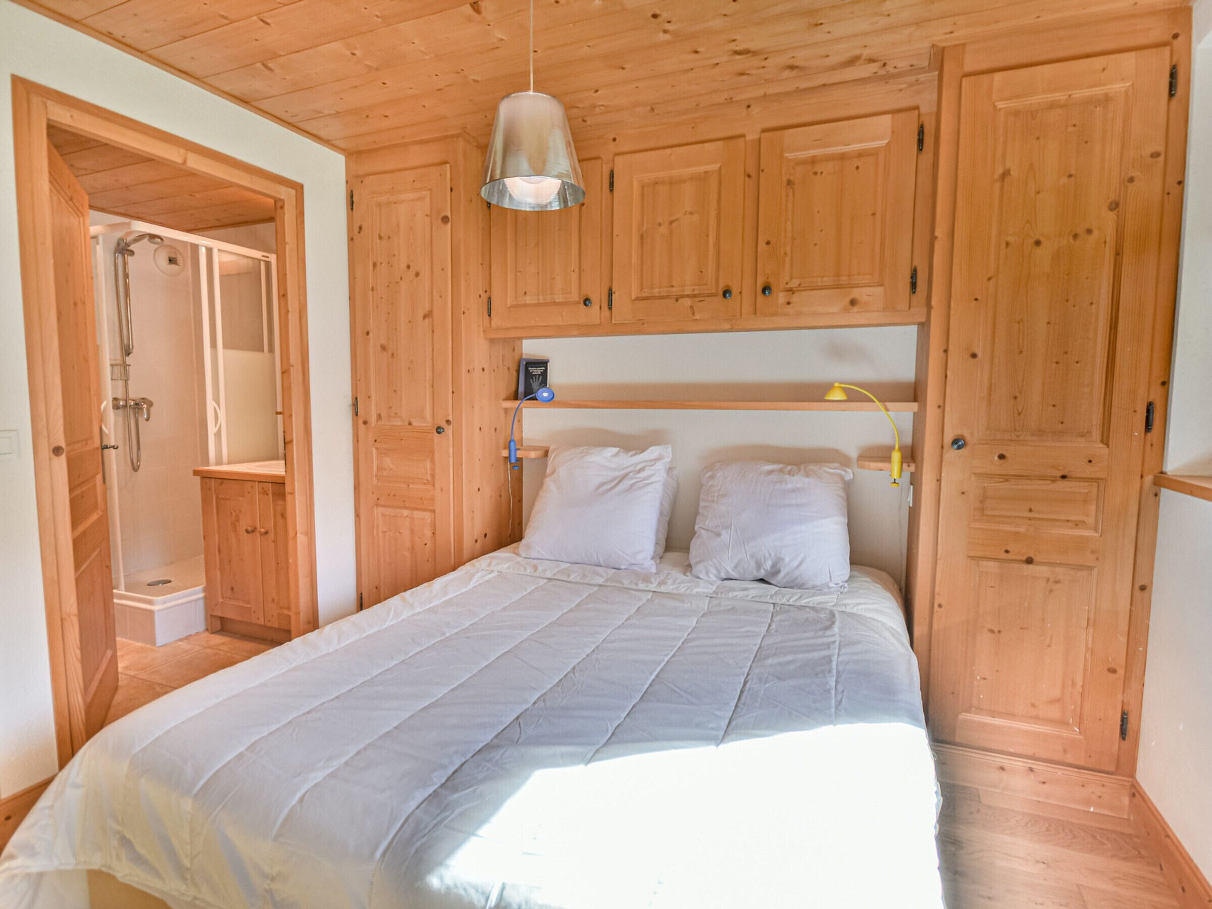 Un dormitorio con techo de madera, una cama con ropa blanca, una mesita de noche con lámpara y una puerta de madera.