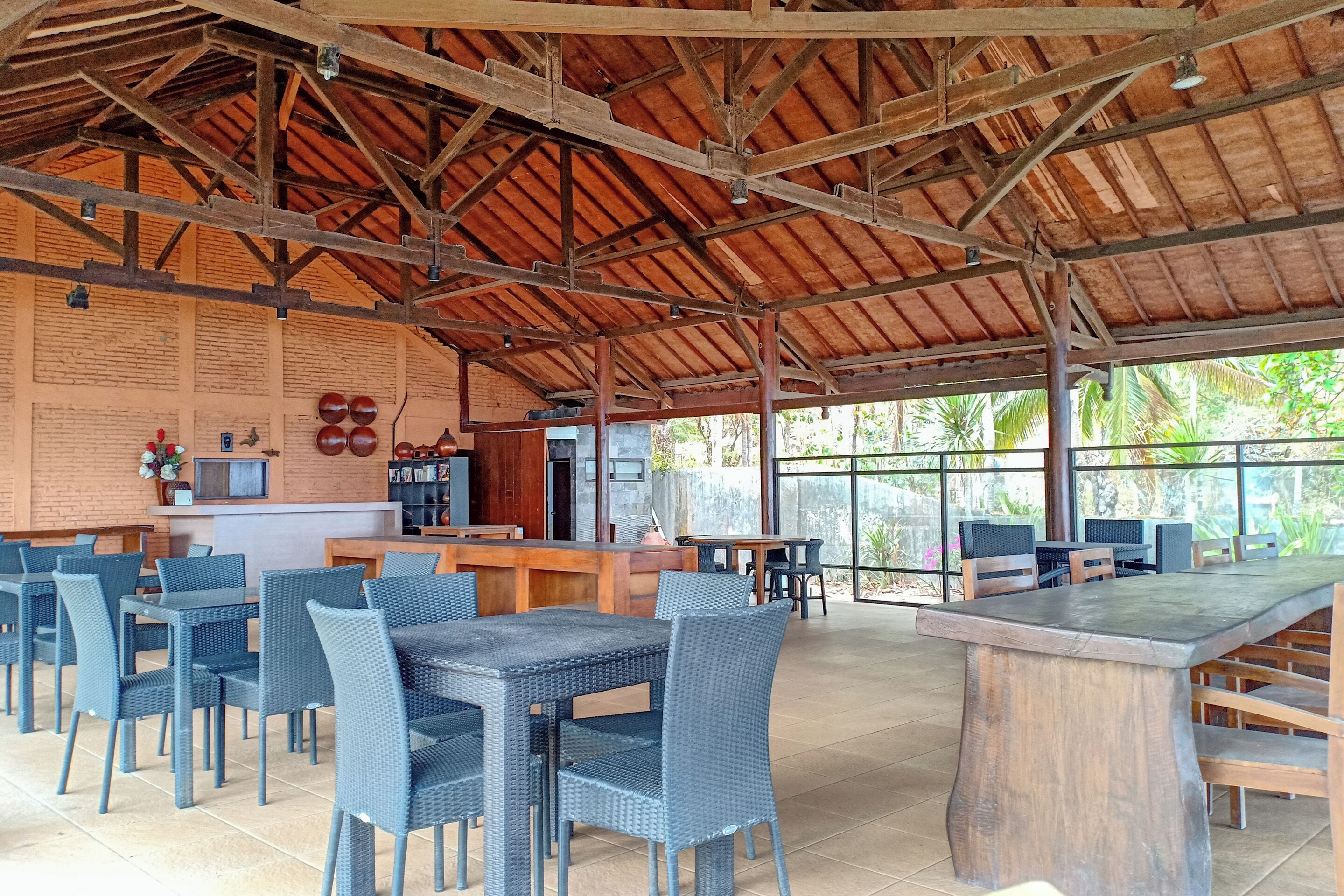 A spacious outdoor dining area with wicker chairs and tables, a wooden bar, and a view of greenery through large windows.