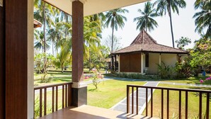 A covered outdoor area with a wooden deck, a view of a garden with palm trees, and a small gazebo. - Mascot Beach Hotel Senggigi (Senggigi)