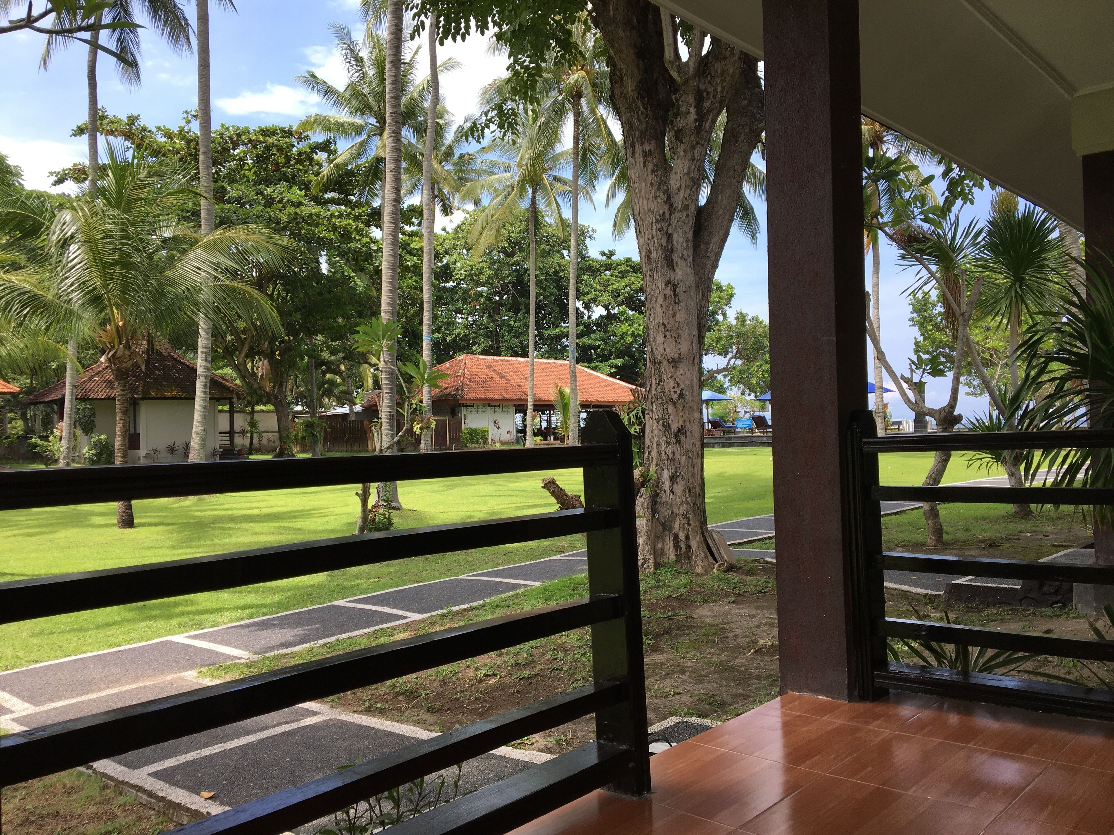 A view from a covered area with a wooden railing, overlooking a well-maintained lawn with palm trees and a building with a red roof.