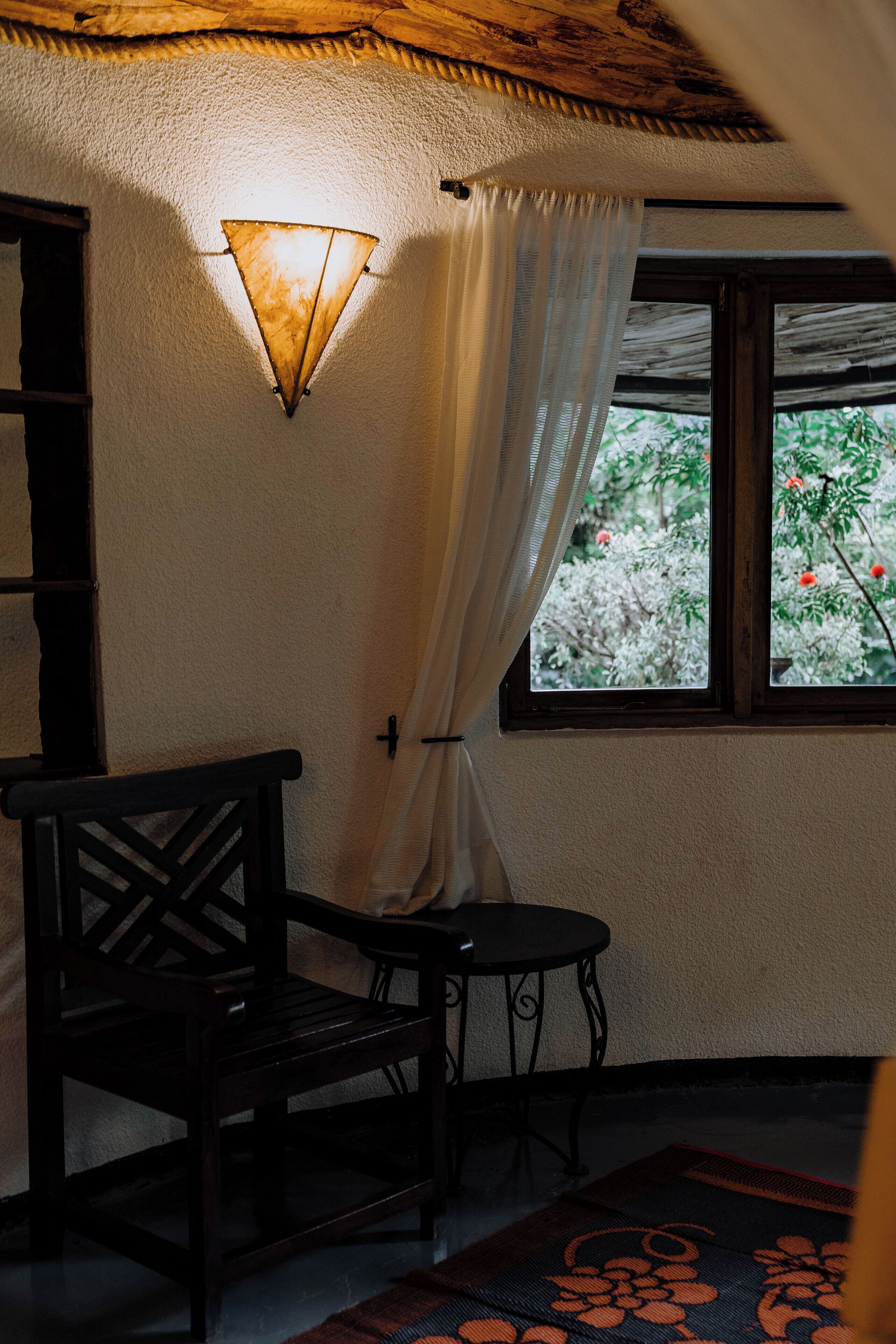 A room with a wooden chair and table, a window with white curtains, and a wall-mounted light fixture.