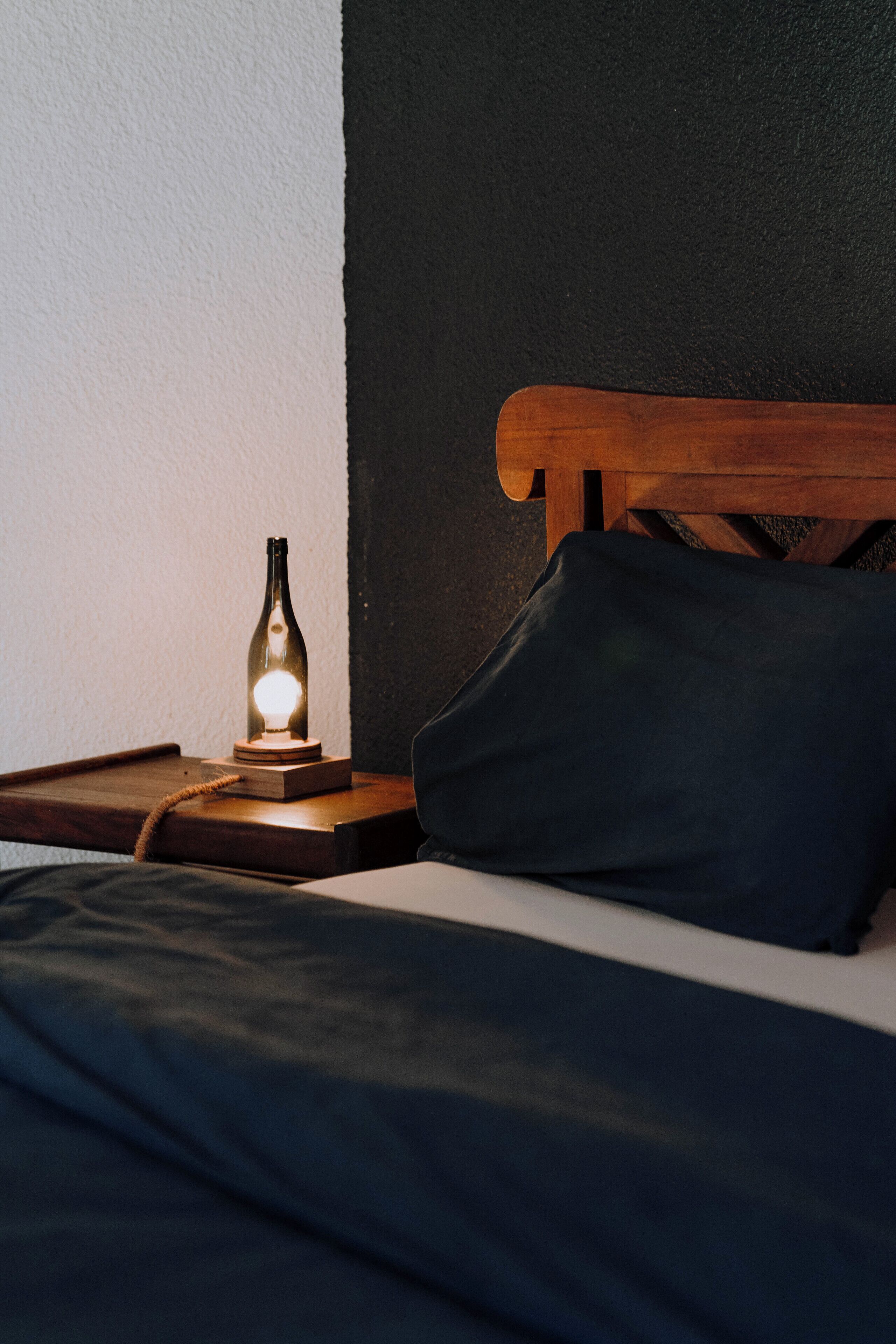 Un lit avec une tête de lit en bois, un oreiller foncé et une bouteille illuminée posée sur un sous-verre en bois.