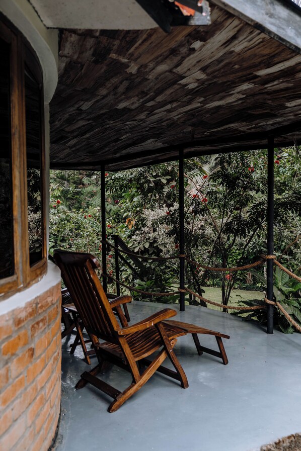 A wooden rocking chair and a brick wall under a covered patio with a view of lush greenery. - Sanna Eco Lodge (Arusha)