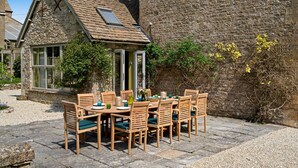 An outdoor dining area with a stone table and wooden chairs, set against a stone wall with climbing plants.