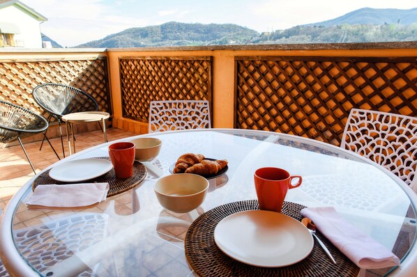A round glass table set for breakfast with plates, bowls, and croissants, surrounded by wicker chairs, on a terrace with a view of mountains. - Casarico (Maslianico)