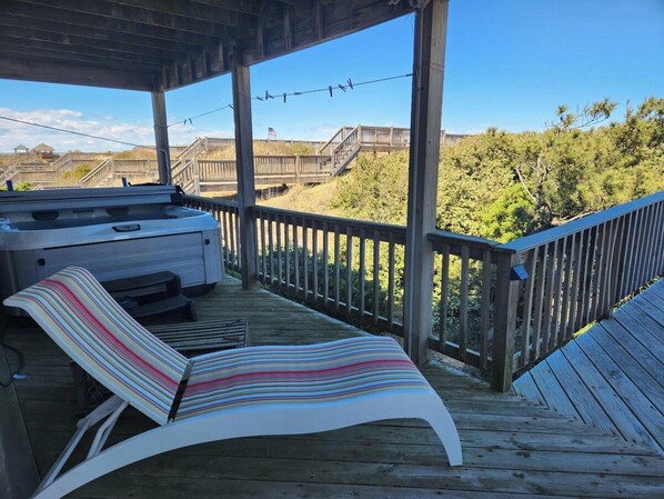 Et overdækket udendørs område med grill, en stribet liggestol og en træterrasse med udsigt over en strand med en trægang og et flag.