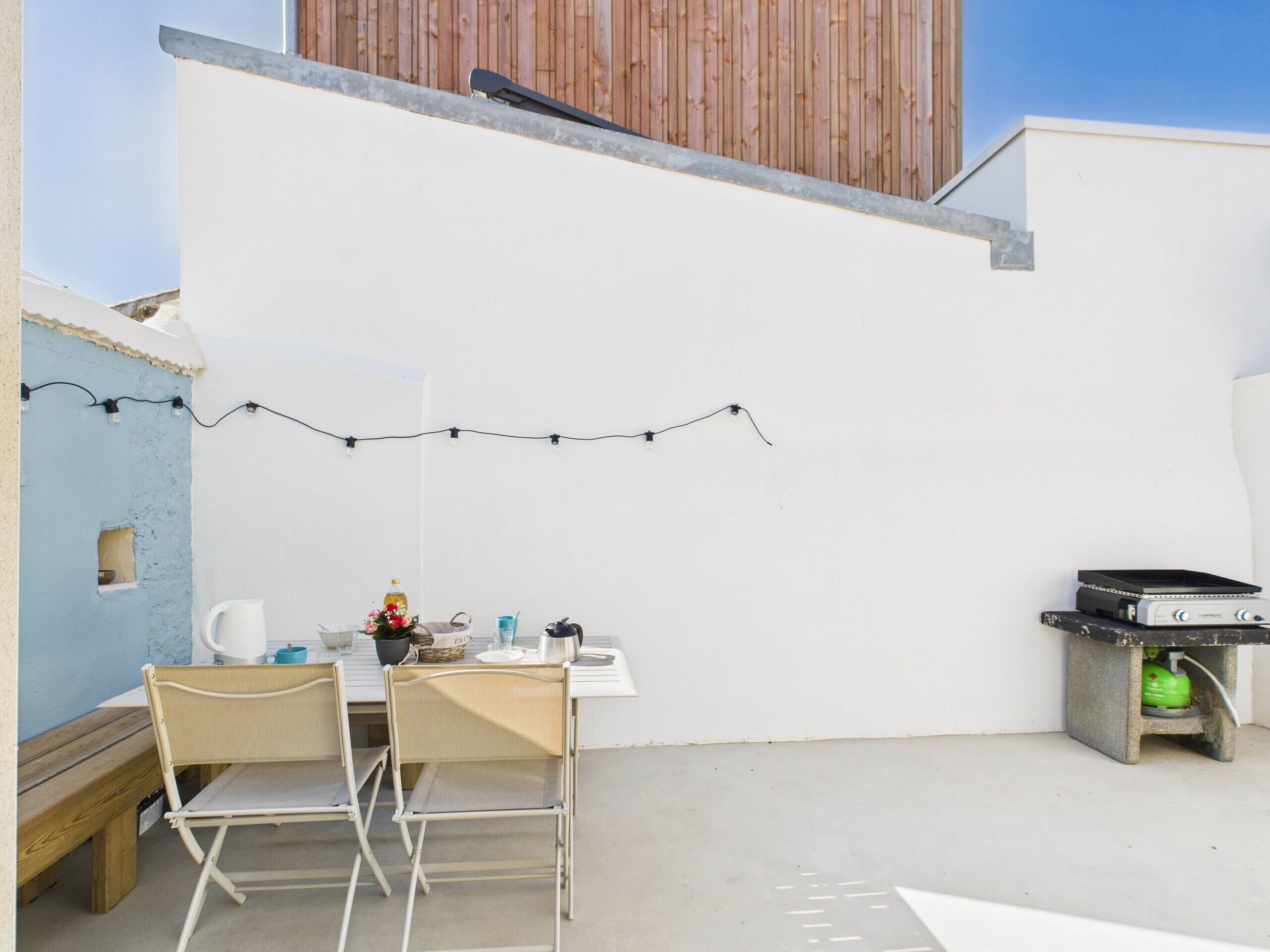 Une petite terrasse extérieure avec une table et des chaises, une guirlande lumineuse, un petit barbecue et un banc en bois.