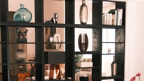 A modern living room with a black sectional sofa, a black ottoman, and a coffee table. A large black bookshelf filled with various decorative items and books is in the background. - Tropical Oasis w pool, AC 10 mins from beach and Brickell (Biscayne Park)