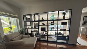 A living room with a beige sofa, a wooden floor, a bookshelf with various decorative items, and a window with blinds. - Tropical Oasis w pool, AC 10 mins from beach and Brickell (Biscayne Park)