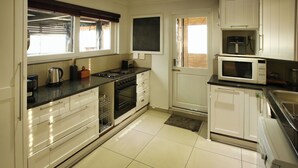 A kitchen with white cabinets, a black countertop, a stove, a microwave, and a window with blinds. - Klopdisselboom Langebaan 10-sleeper (Langebaan)