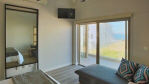 Ein modernes Hotelzimmer mit einem großen Bett, einer Couch mit gemusterten Kissen, einem kleinen Tisch und einem großen Fenster mit Blick aufs Meer.