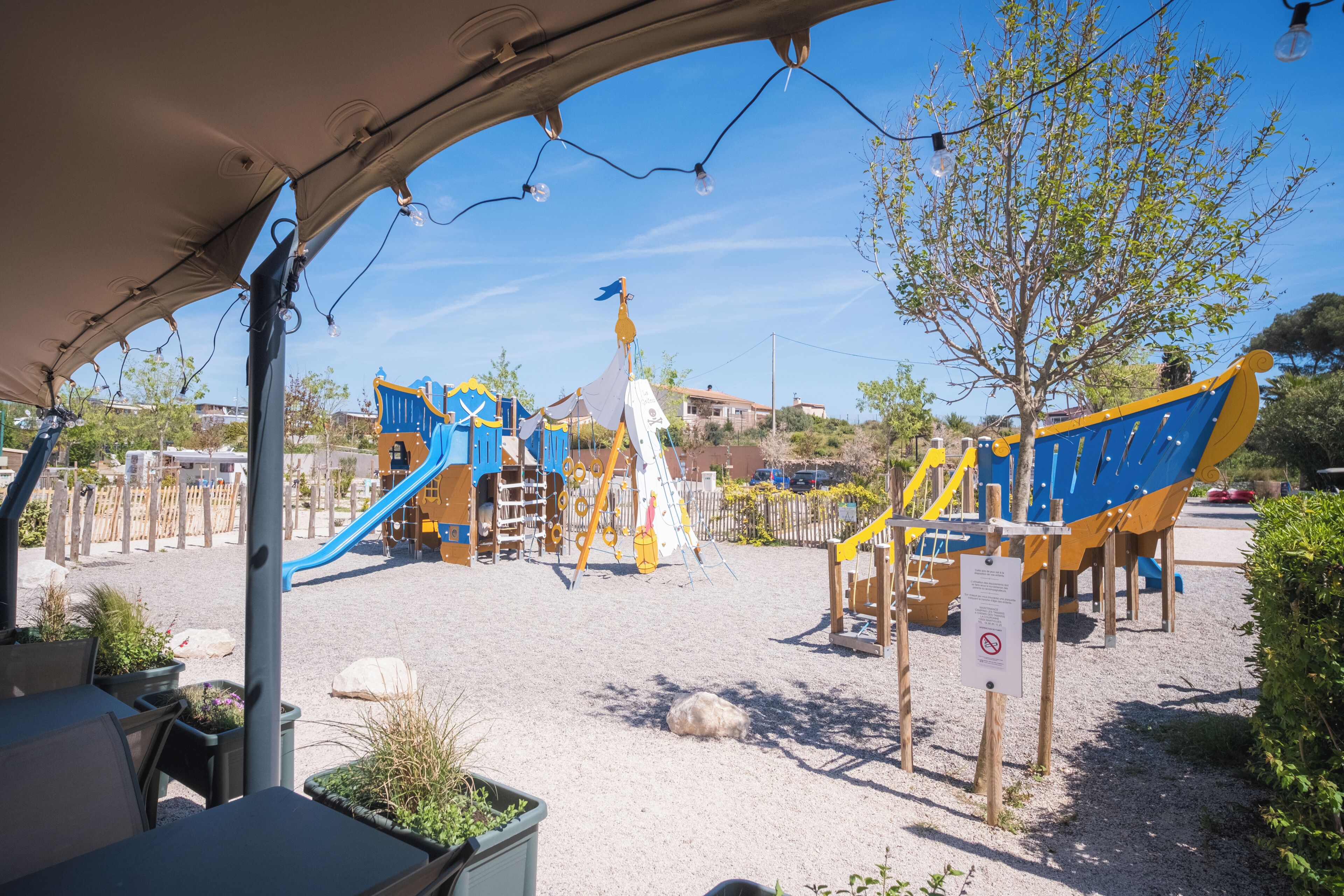 A playground with slides and climbing structures, surrounded by a sandy area and some greenery.
