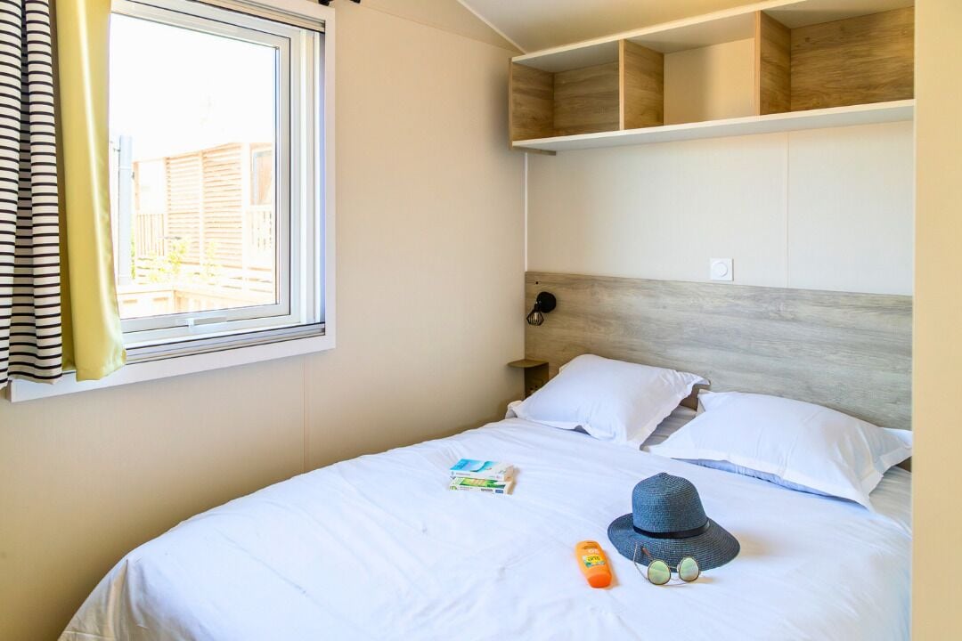 A hotel room with a bed, white bedding, a window with curtains, a bedside lamp, and a hat on the bed.