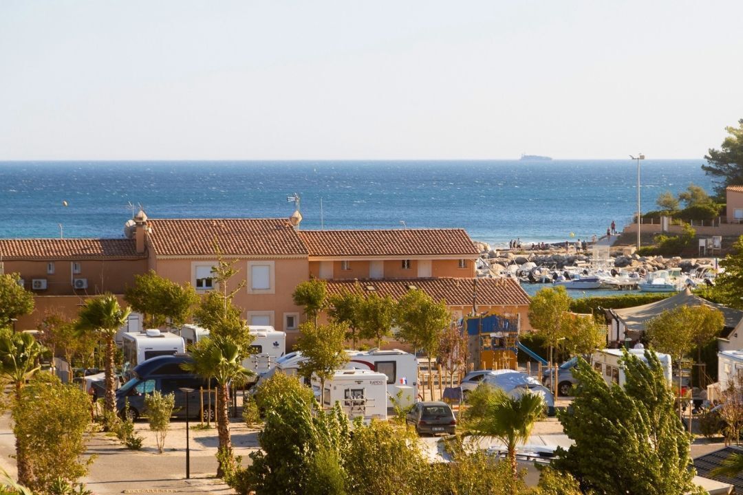 A coastal area with a row of buildings, some with balconies, and a marina with boats. There are RVs parked in front of the buildings.