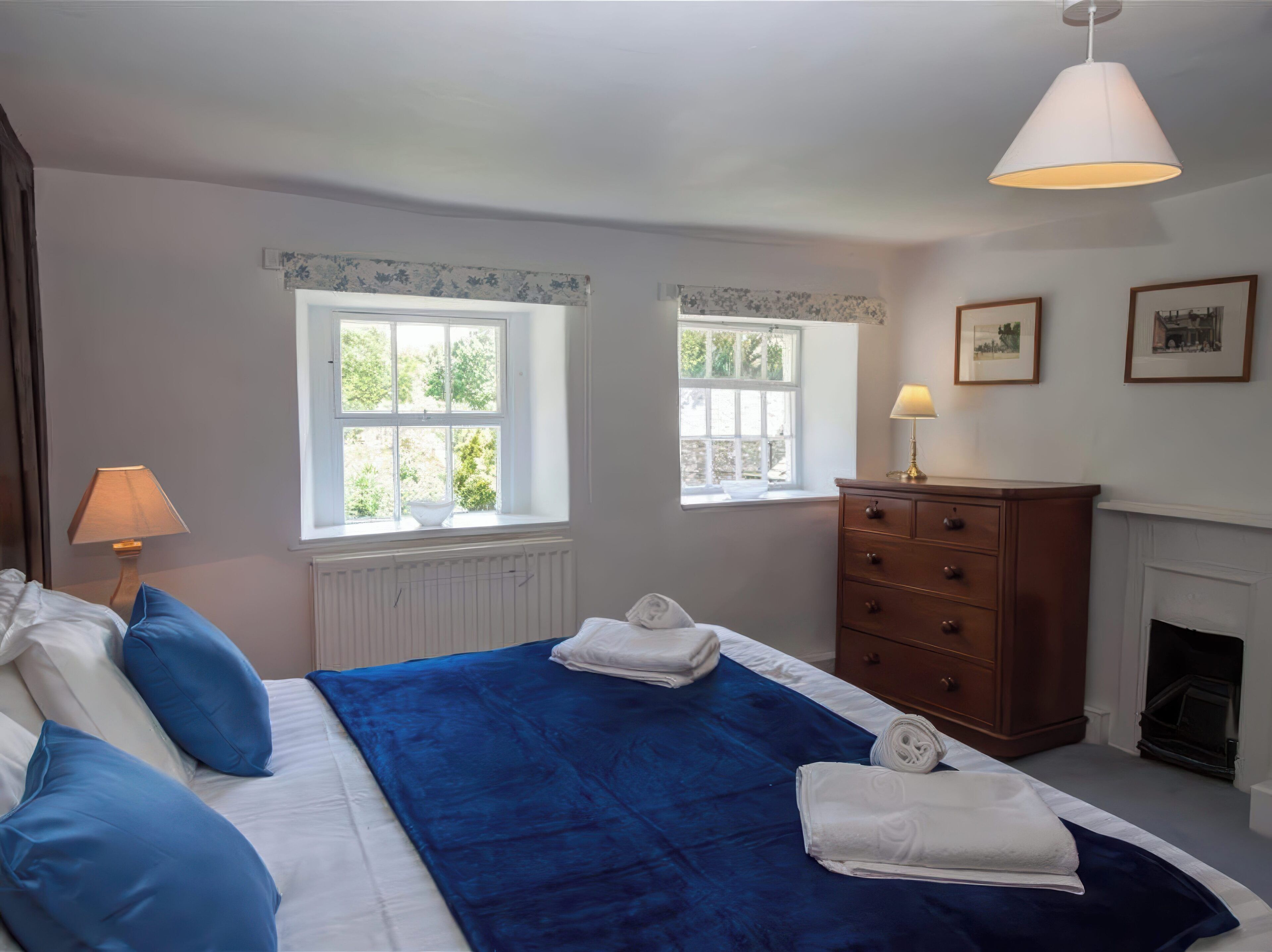 A bedroom with two windows, a bed with blue bedding, a wooden dresser, and a white lamp.