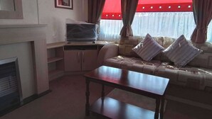 A living room with a sofa, a table, a television on a stand, and a fireplace. - Sleep4 Cabin - Self-checkin, Free Parking, Petok (Lowestoft)