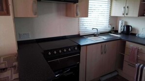 A compact kitchen with a stove, oven, sink, and a window with blinds. - Sleep4 Cabin - Self-checkin, Free Parking, Petok (Lowestoft)